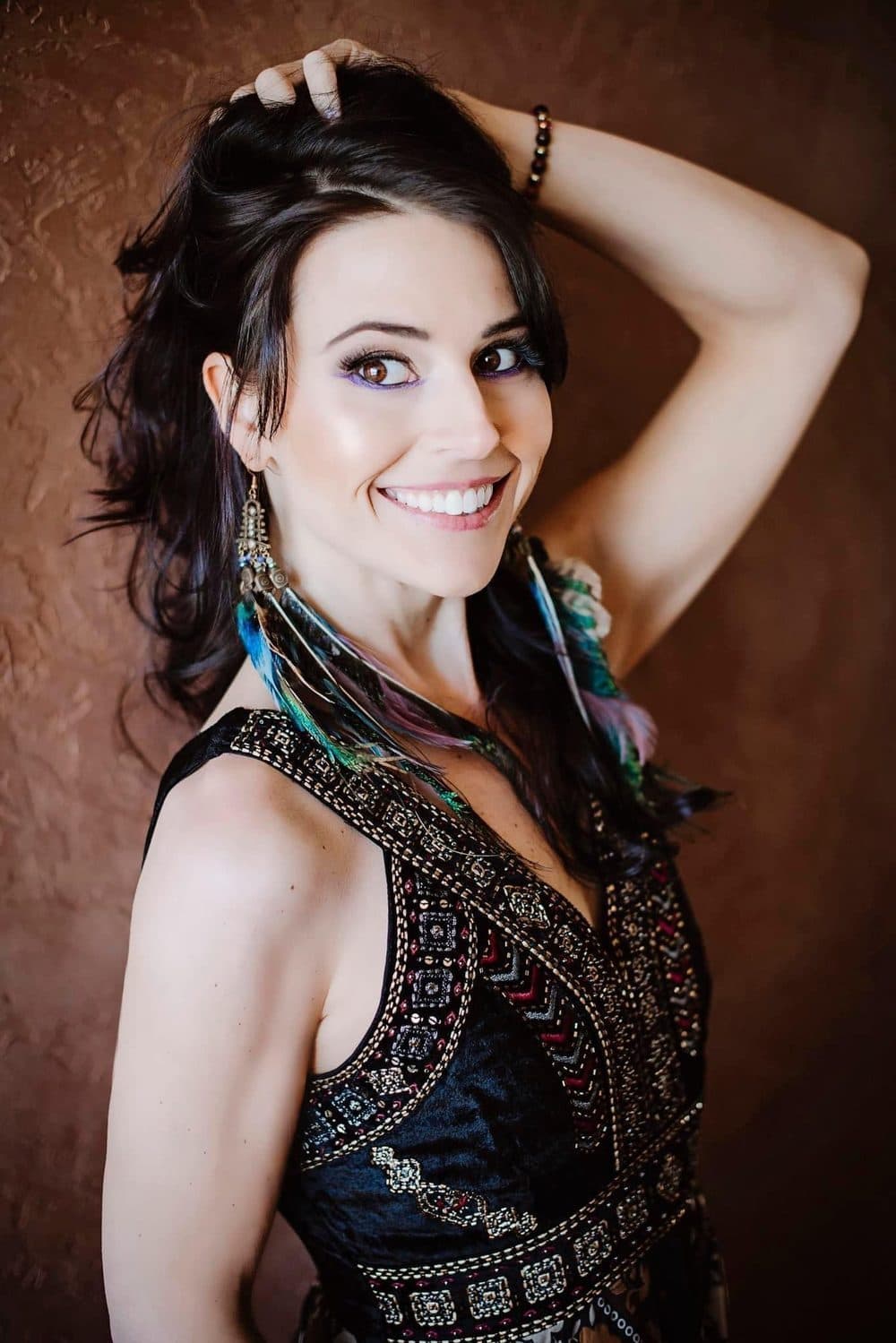 Solara Rose laughing with one hand in her dark hair, wearing an embroidered dress and feather earrings against a terracotta wall