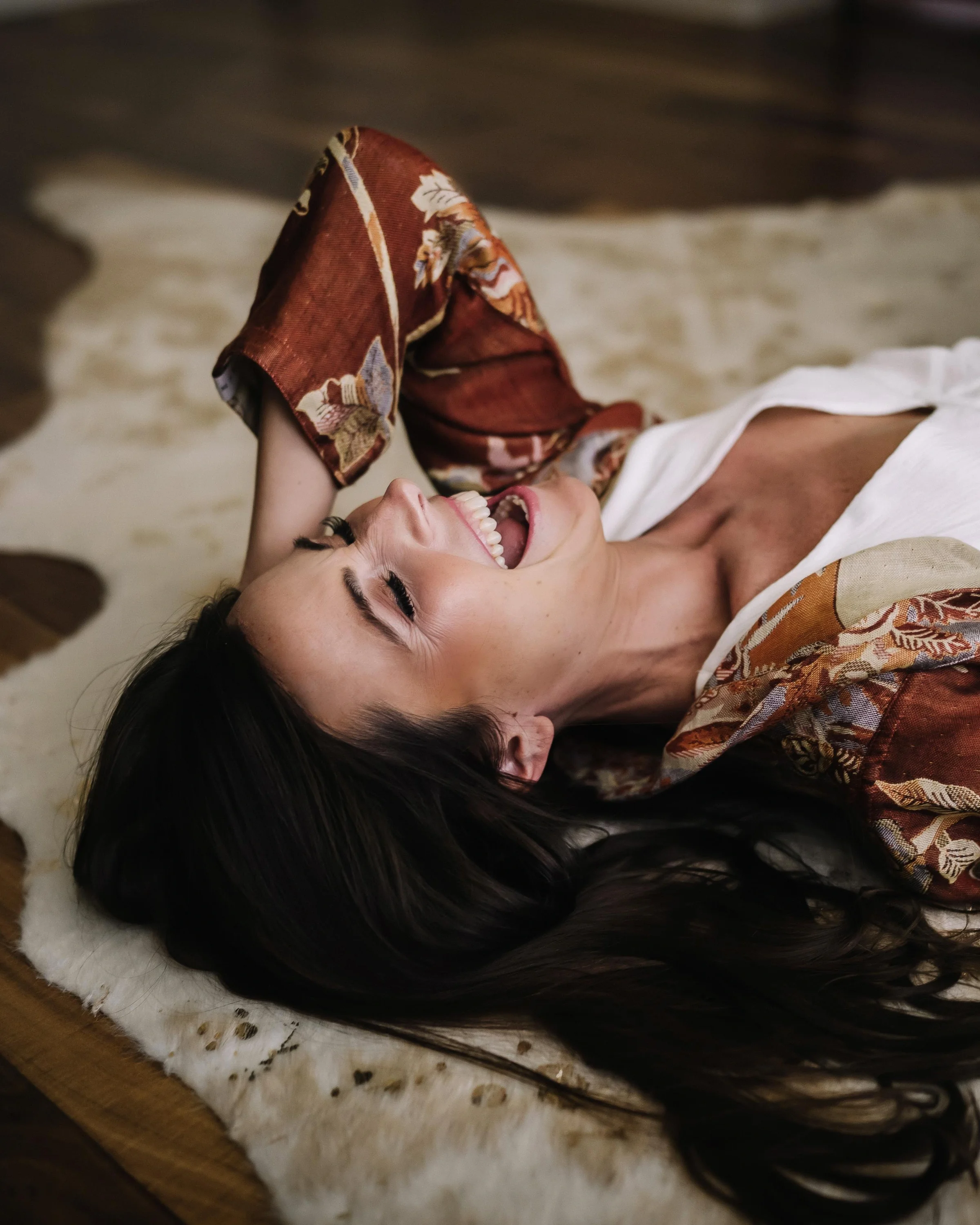 Solara Rose laughing with joy, lying back on a cowhide rug in a white top and patterned robe